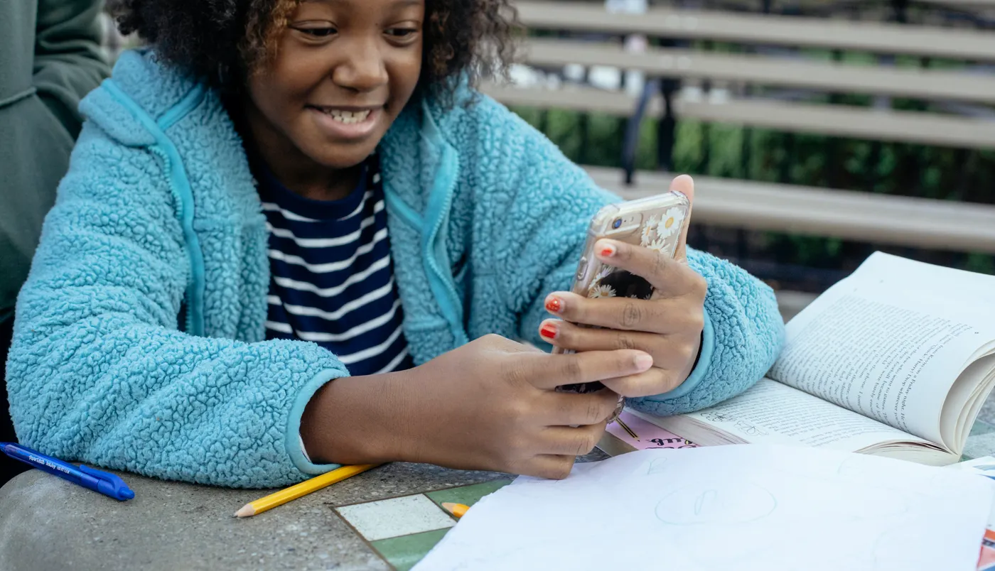 A child smiles at a phone in her hands, with schoolbooks open on the table