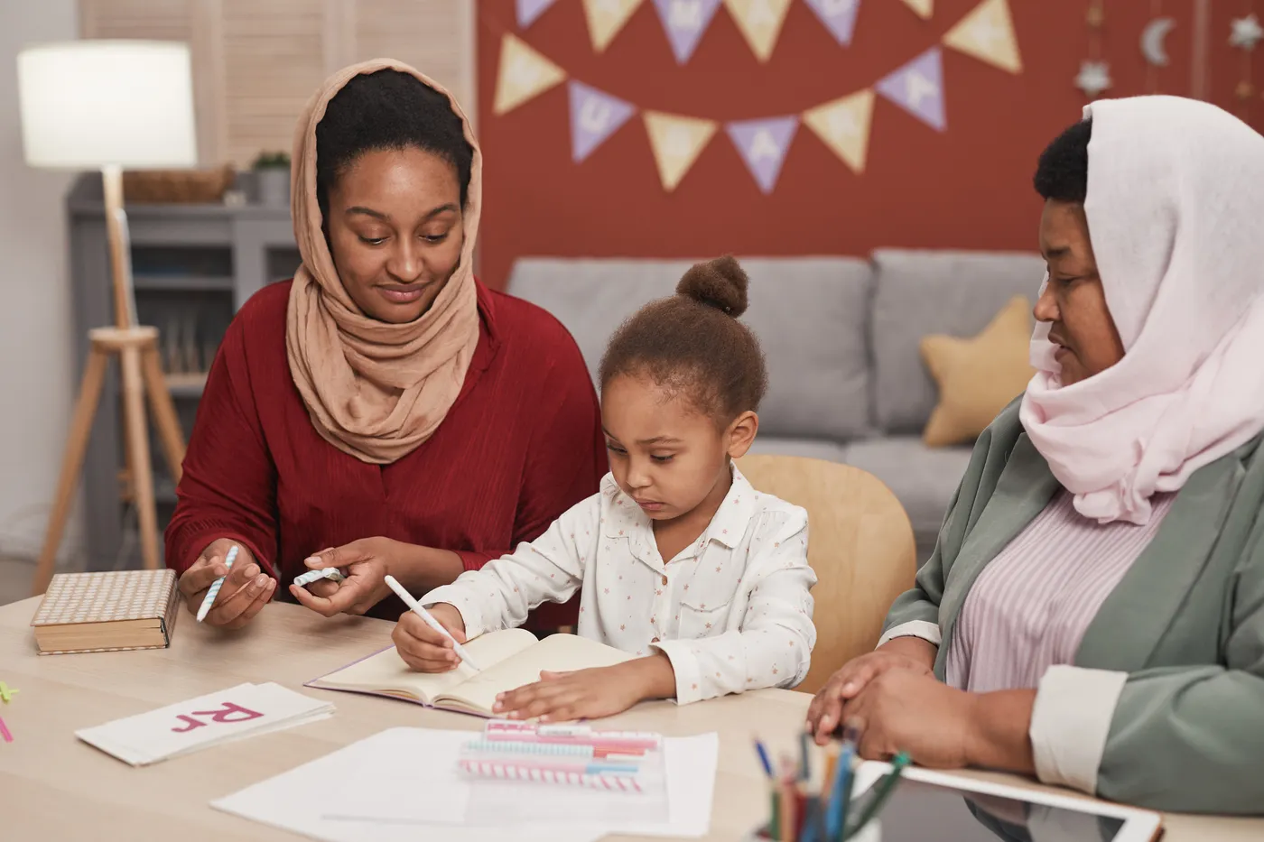 A young child practises writing at a table with her mother and grandmother beside her