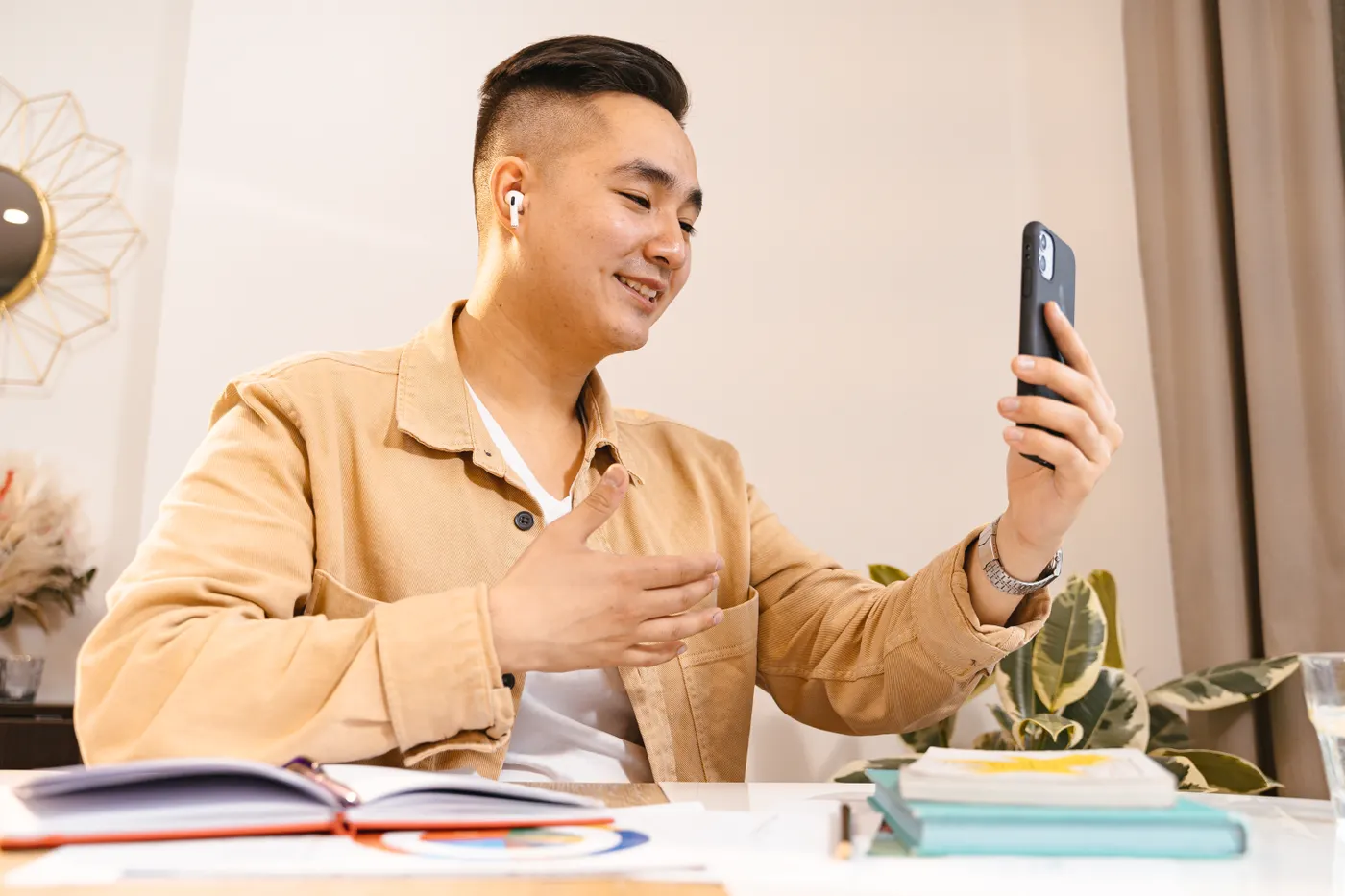 A tutor wearing earbuds talks to a student on a phone held in his hand, with books on the desk